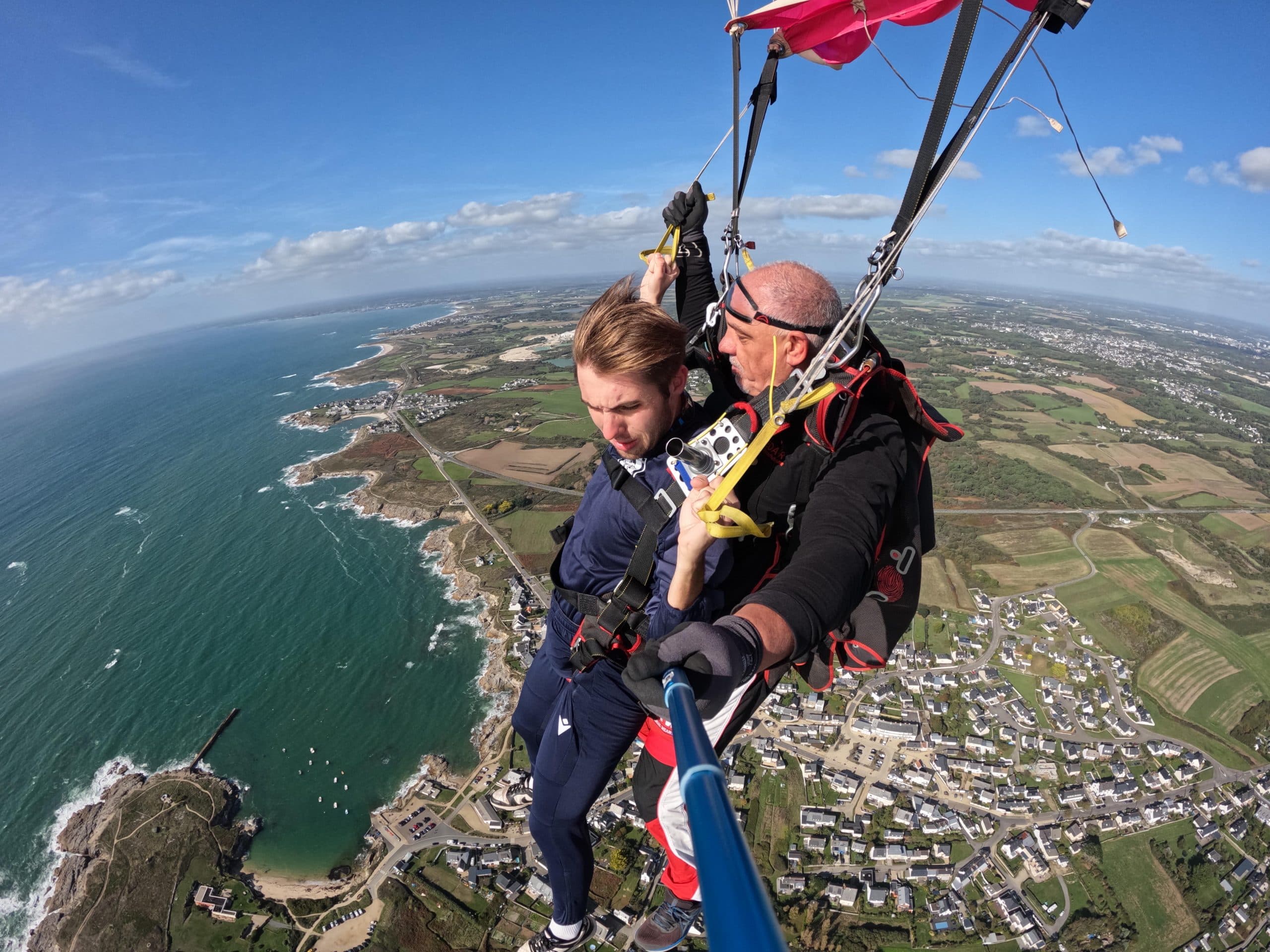 Jeune homme en saut tandem sous parachute rouge avec moniteur, vue sur village côtier breton et côte rocheuse de Péréllo – Saut en parachute Bretagne