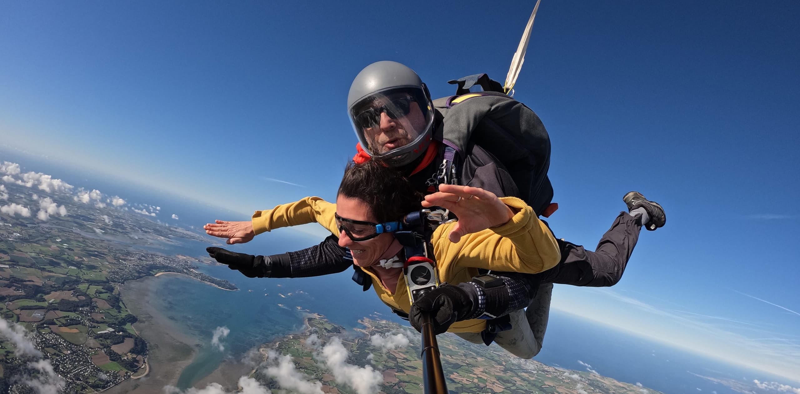 Femme souriante en veste jaune bras écartés en chute libre tandem avec moniteur casqué, au-dessus de la baie de Morlaix sous un ciel bleu dégagé – Saut en parachute Bretagne