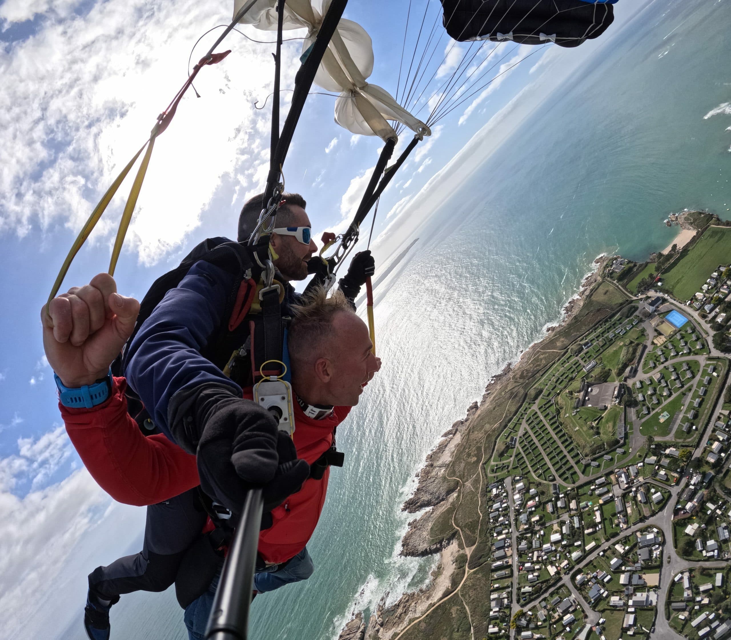 Homme poing levé lors de l'ouverture du parachute en tandem au-dessus du village côtier de Péréllo, moniteur à lunettes de soleil, mer turquoise et falaises en arrière-plan – Saut en parachute Bretagne